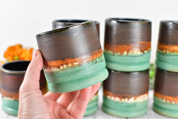 hand holding a handmade wheel thrown ceramic pottery whiskey tumbler cup in brown, bronze and turquoise patina green glaze by artist Camille Woodward