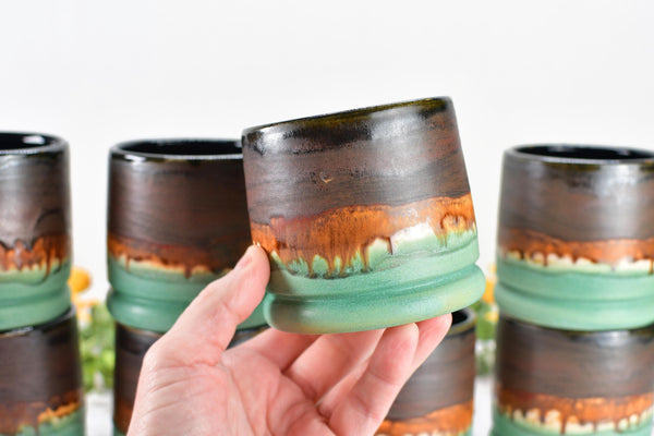 hand holding handmade wheel thrown ceramic pottery whiskey tumbler cup in brown, bronze and turquoise patina green glaze by artist Camille Woodward