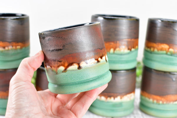 hand holding handmade wheel thrown ceramic pottery whiskey tumbler cup in brown, bronze and turquoise patina green glaze by artist Camille Woodward