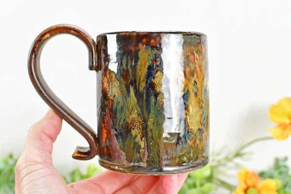 Hand holding a bronze, tan and Black Coffee Mug with Drippy, Earthy Ceramic dhiny glazes. Pottery Cup Handmade by Camille Woodward artist.