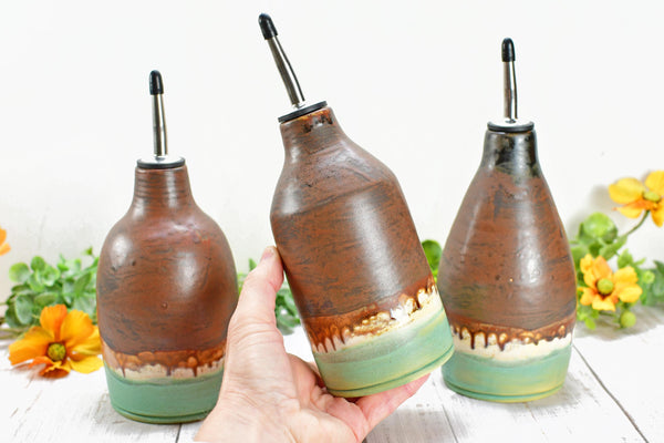 Hand holding one Ceramic Olive Oil Dispenser. Three Handmade Pottery Liquid Soap Bottles in Stoneware Bronze, Copper, Rust, Turquoise glazes for Bathroom and Kitchen Decor. Camille Woodward artist.