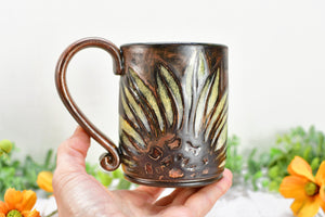 Hand holding a copper and yellow stoneware, water etched sunflower coffee mug with orange flowers in the background.