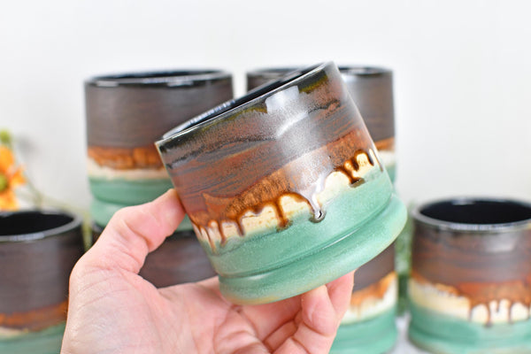 May include: A ceramic mug featuring a dark brown upper section that transitions into a lighter cream band, finished with a sage green base. The glaze displays a distinct drip pattern where the brown glaze runs down over the cream and green layers.