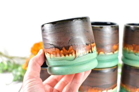 Hand holding a handmade wheel thrown ceramic pottery whiskey tumbler cup in brown, bronze and turquoise patina green glaze by artist Camille Woodward 